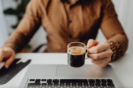 Adulto de camisa xadrez segura uma xícara de café sobre o teclado do notebook em uma mesa de trabalho