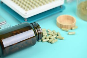 Frasco âmbar deitado com cápsulas verdes de nutracêuticos e máquina de enchimento de cápsulas ao fundo
